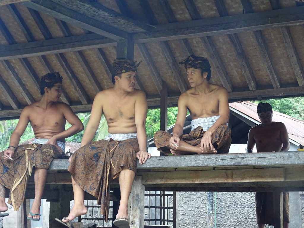 There are three men sitting on the edge of a roofed area talking to each and there is a fourth men on the back doing something that is not visible on the photos. The men are wearing a traditional festive Indonesian brown colored skirt and they are wearing head covering brown scarfs