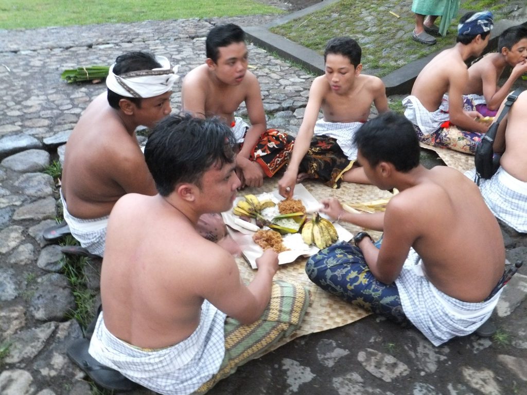 A group of five men sitting on the floor and wearing only a skirt. They are sharing a dish of traditional food before starting the fight.