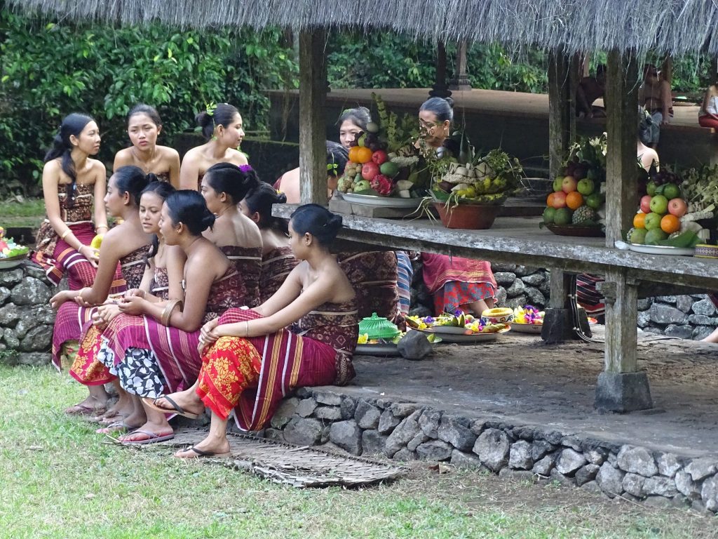 A group of ten women dressed up with the traditional Indonesian dress and sitting on the sacred space on Tenganan waiting for the ritual to start. They are sitting on the stair of a bale. There are several trays with fruit on the bale to be used as offerings during the festival