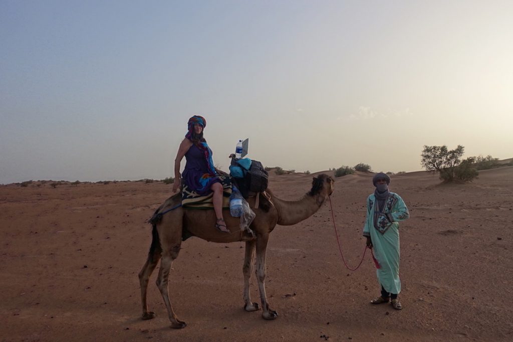 Pilar riding a dromedary with a tour guide on the Sahara desert at sunset time during the Zagora desert tour to Erg Lihoudi