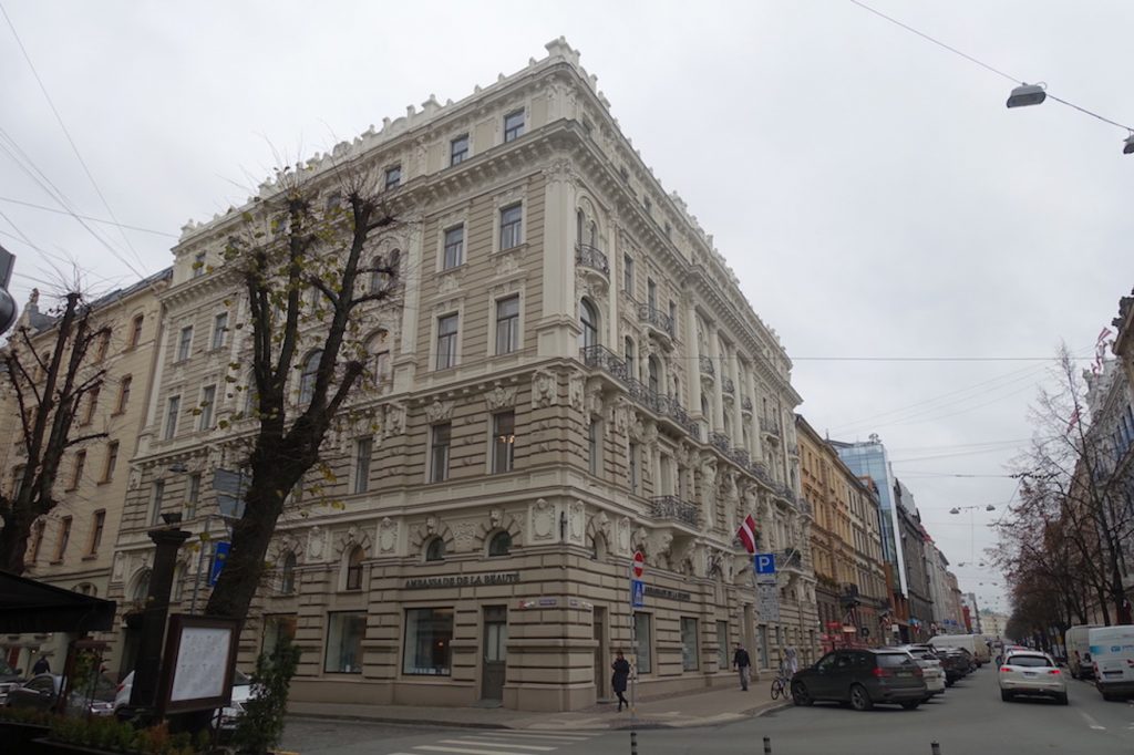 The corner of an Art Nouveau building in Riga. The color of the building is light grey and there are two trees on the left of the photo. On the right of the photo you can see a line of trees and a street with some cars parked on it and a man walking.