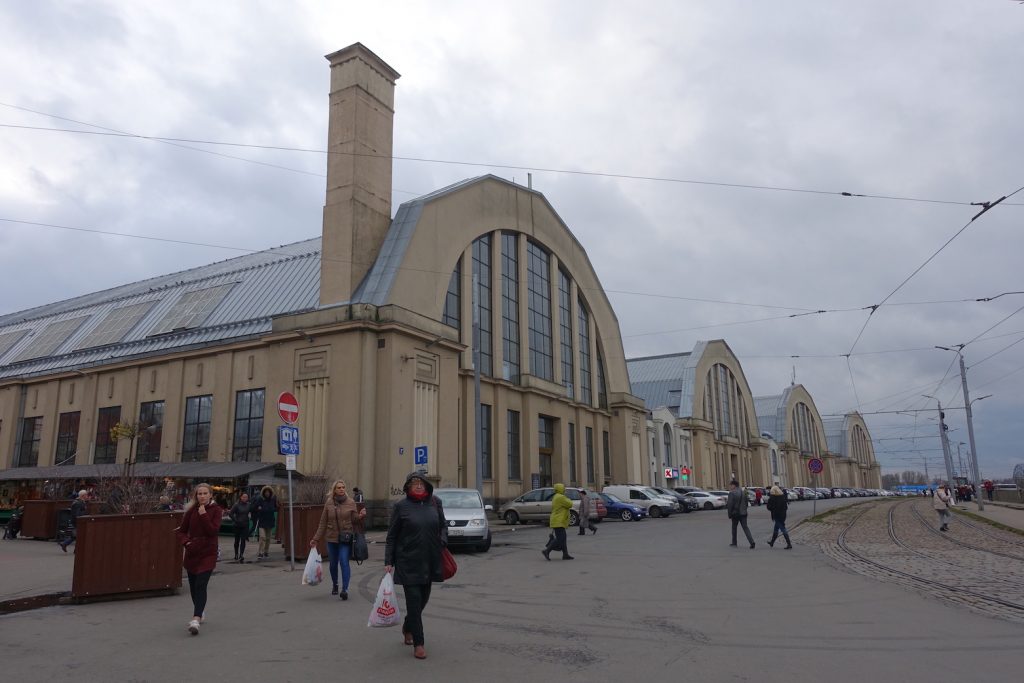 This is a photo of the Riga central market from outside There are four round arc shaped buildings. The color of the buildings is yellow sand  with blue round roofs. There are cars parked outside and people walking.