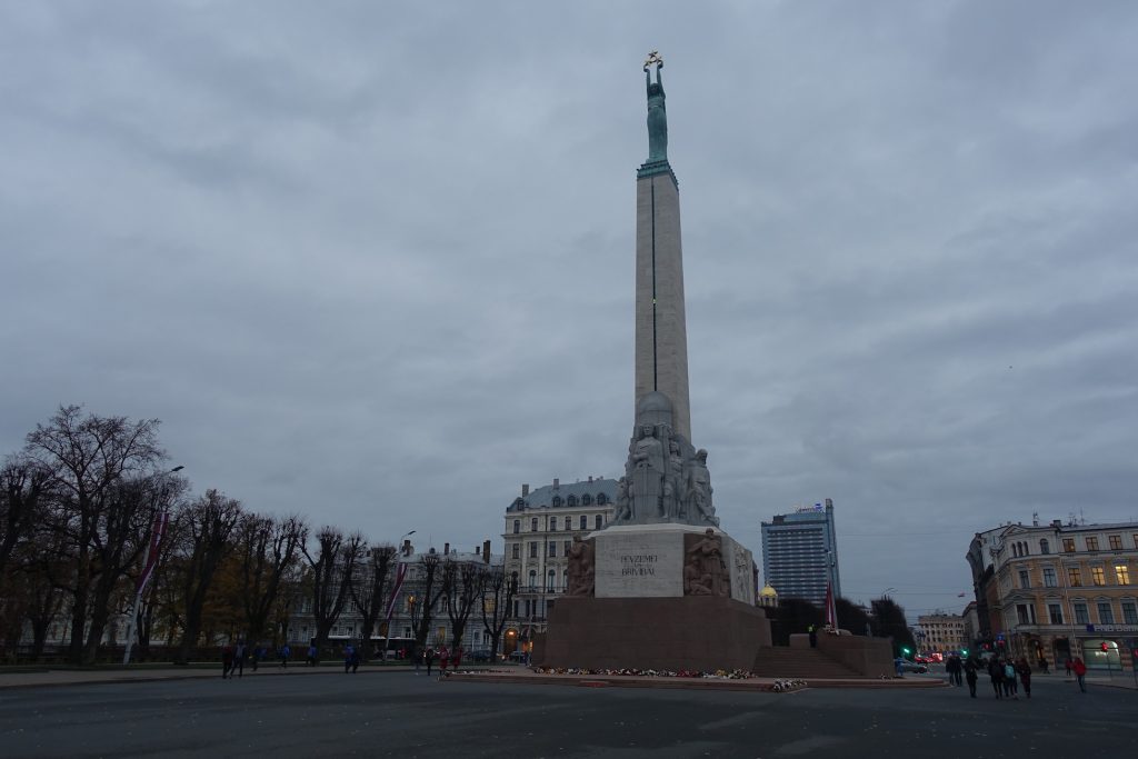 Freedom monument. It is a rectangular tower standing on its own with a statue on the top. There is a line of tress on the left and some buildings on the back.