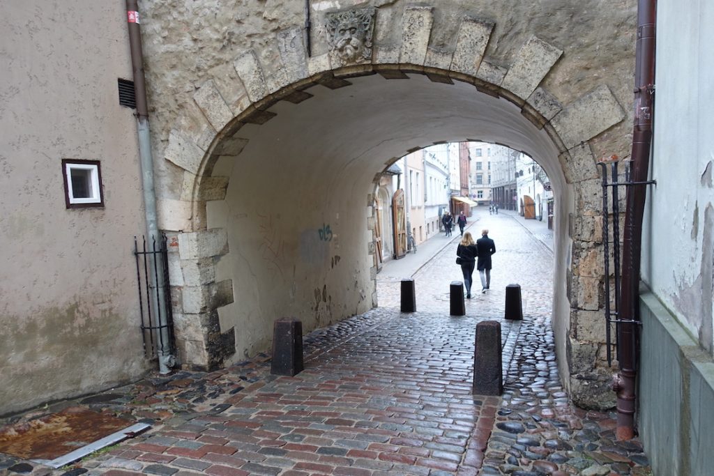 Riga Swedish gate. It is an arc and under the arc you can see a street where there is a couple walking. The street floor is wet as it has been raining.