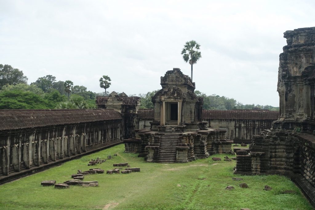Angkor Wat main temple. You can see several buildings with columns and statues, A section with grass and some trees on the background.