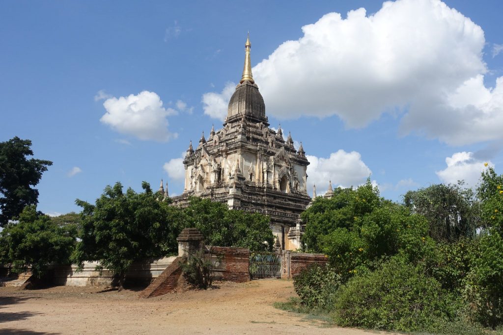 The Gawdawpali temple with its golden top spire and its grey dome. The temple is surrounded by trees and there are some clouds on the blue sky