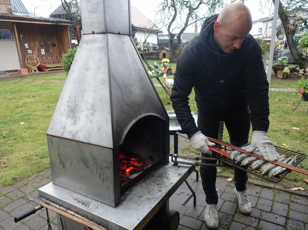 Man grilling Lamprey at Carnivaka garden on a metal over. The man is wearing white gloves and holding a tray with several fish ready to be prepared on the metal oven with burning coal.