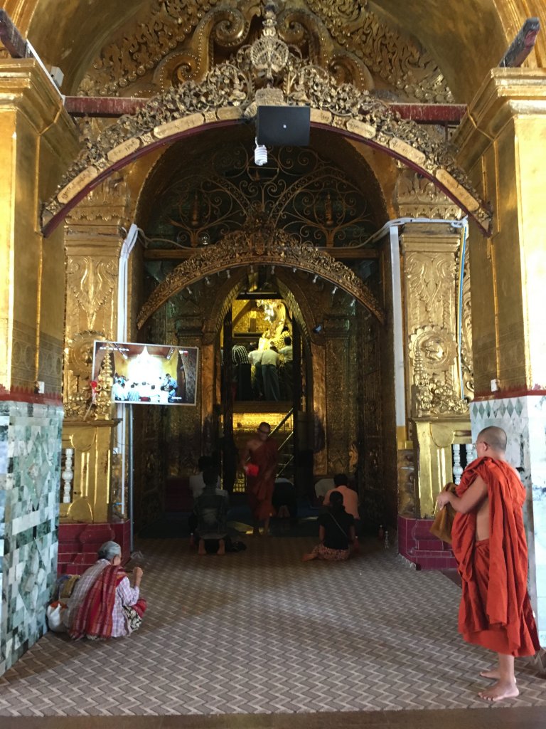 Some people inside the Mahamuni pagoda watching the ceremony on the T.V screens. There is a Buddhist monk dressed in orange on the right hand side