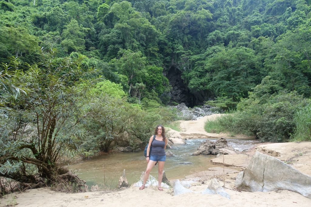 Pilar at the river side on the hike heading to Tra Ang cave. The river is pretty dry as it is dry season and there are a lot of tree. Pilar is wearing shorts and a grey tank top