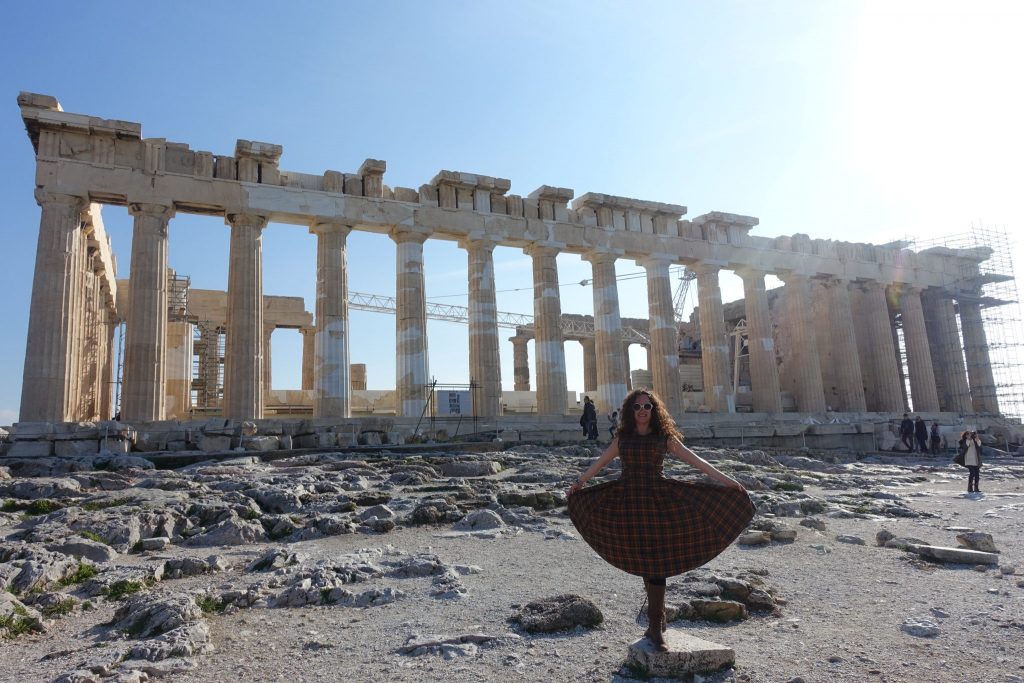 Pilar wearing a dress and expanding the dress with her hands and the Parthenon in the Acropolis, in the back. The Parthenon is one of the most iconic Athens landmarks