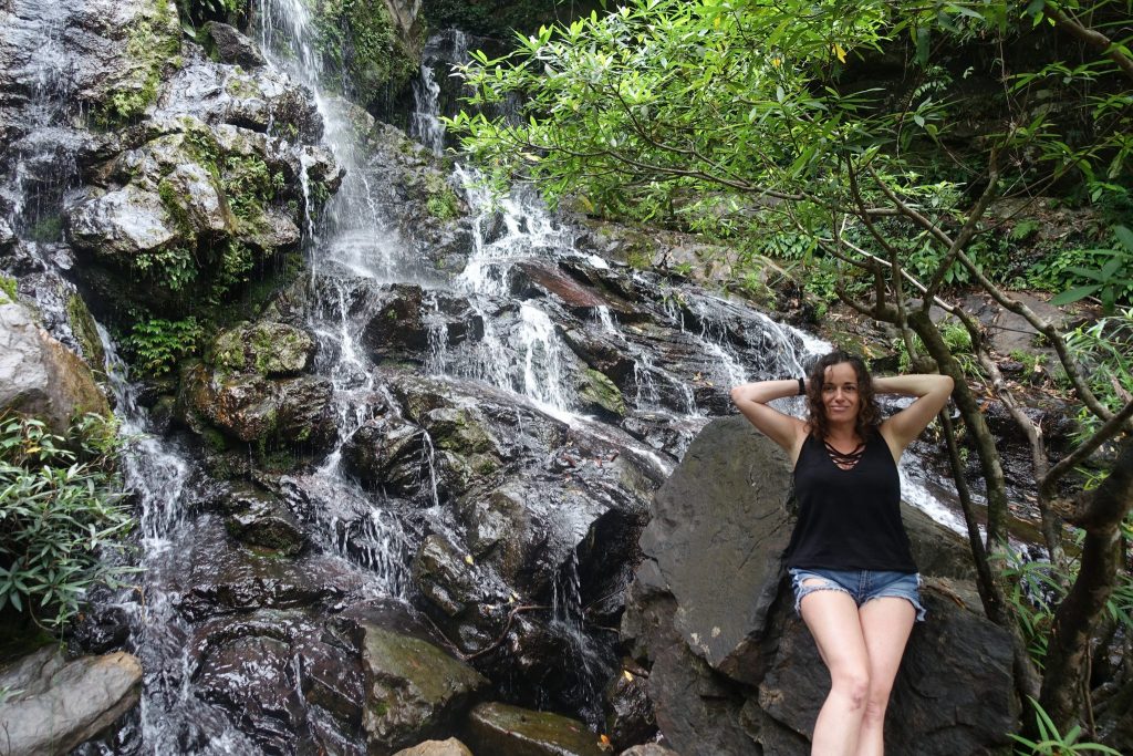 Pilar posing leaning on a rock with a waterfall behingdand a tree on  the right hand side. In the Phong Nha botanical gardens