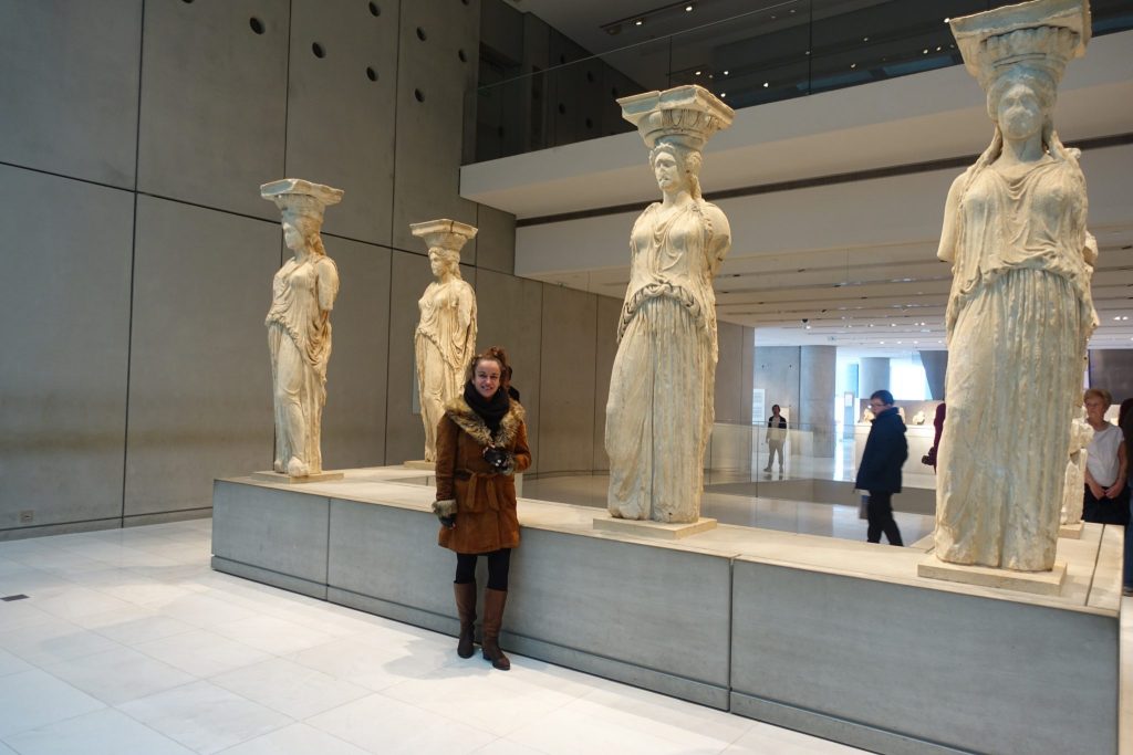Four woman columns, caryatids in the interior of the acropolis museum. Pilar is posing with a brown jacket in front of the caryatids.