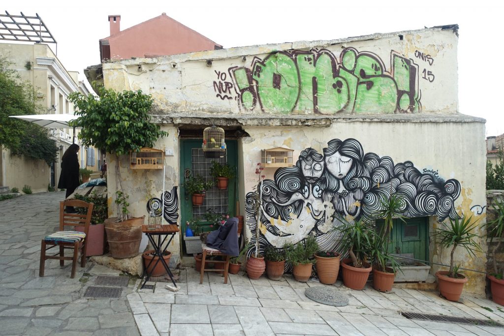 A building with graffities in the Plaka area. The Grafitti consist of some green letter and several heads of women with lot of wavy hair. There are some pots with plants outside of the building and a table and two chairs.