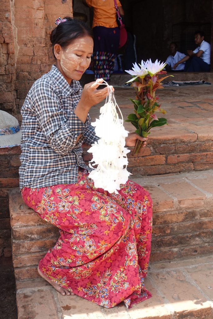 Burmese woman holding some white flowers on both hands and wearing Thanaka on her face. She is selling the flowers at the door of one of the temples.