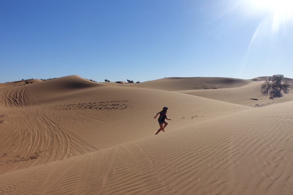 A landscape of sand dunes and Pilar walking up a sand dune, during the Sahara desert tour.