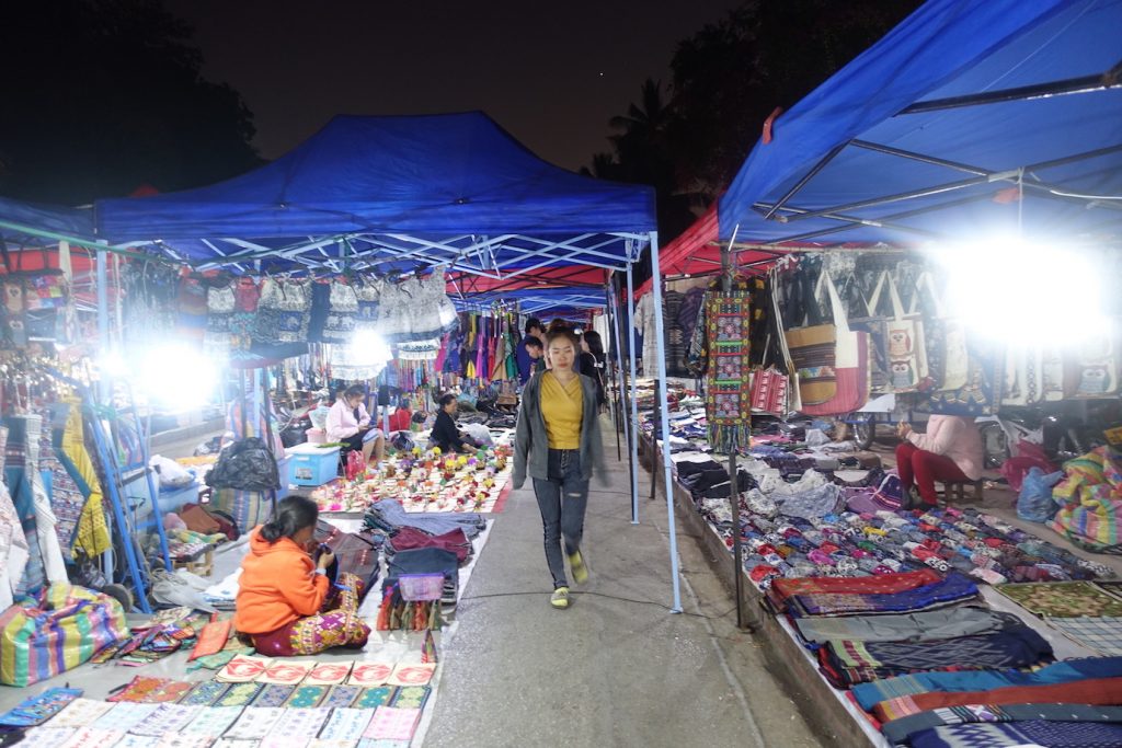 Luang Prabang night market. Some stalls with blue roofs and a women walking toward the person who took the picture. They sell clothes in a variety of colors.