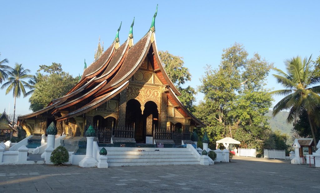 Wat Xieng Thong temple. It has a white stairs entrance and brown roof with green spires, Asian style. There are several trees and palm trees.