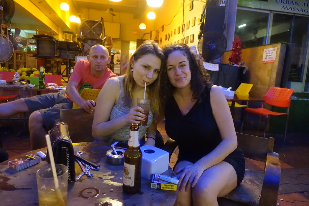 Pilar sitting at a terrace with a blonde girls and a guy. The girl is sipping a cocktail while posing on the photo with Pilar. The photo is taken in Soi Rambutrri street and there are a few beers and cigarrettes at the table