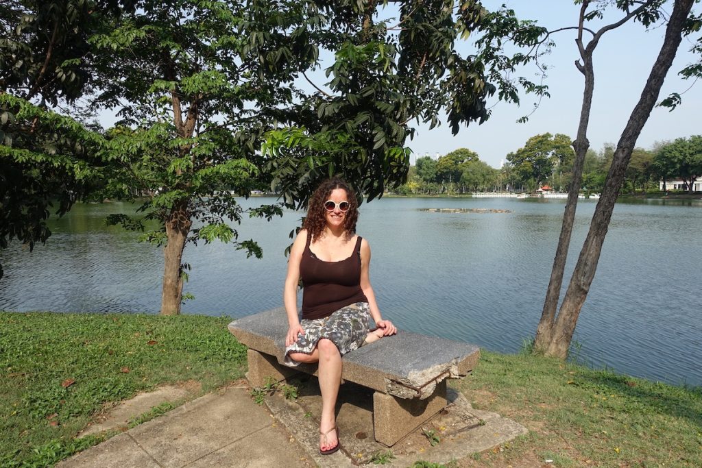 Pilar sitting on a bench in the Lumpini park in Bangkok. There are some trees and a body of water on the abck. Pilar is wearing round sunglasses, a skirt with flowers and a brown top