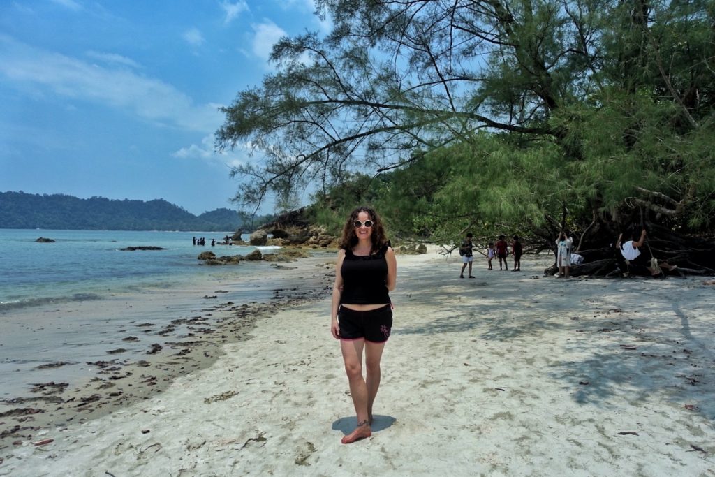 Pilar wearing a black short and sleeveless T shirt at the Buffalo beach. There are some trees and rocks at the beach, and you can see the sea and a nieghbouring island