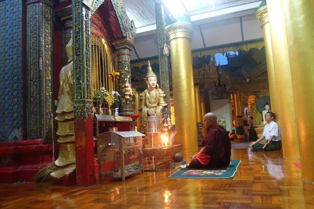 A buddhist monk and a man in a lotues position in front of an altar with some Buddha statues in the Shwedagon pagoda in Yangon Myamar
