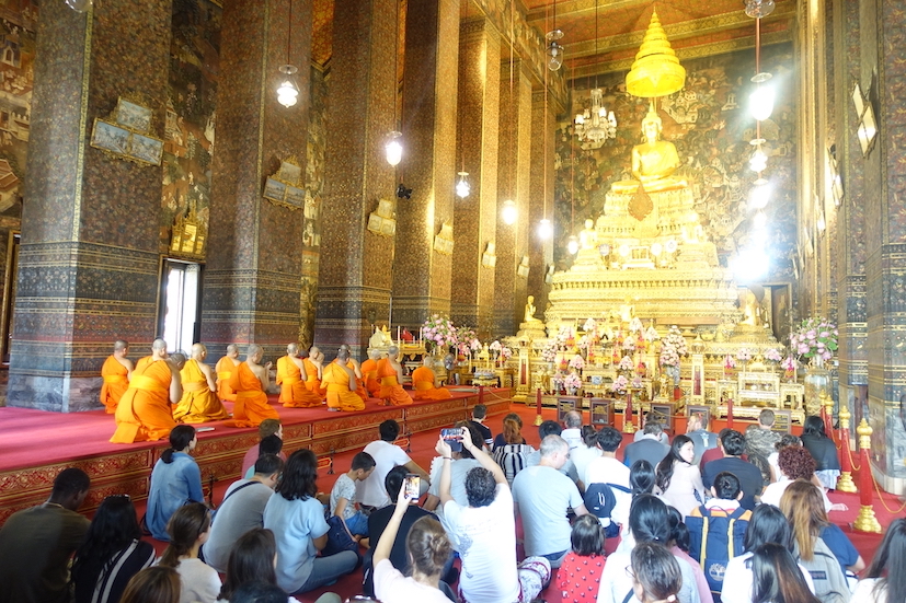 A golden Buddha statue and several Buddhist monks, dressed in orange on the left side of the praying room. One the right hand side some lay poeple praying on their knees. This is in the Wat Pho temple in Bangkok