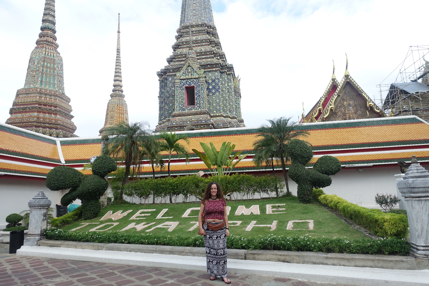 Pilar at the entrance garden of the Wat Pho temple in Bangkok. Wat Phoe is written on the grass with some flowers. There are some palm trees and other bushes and the roof of a building and some chedis on the back