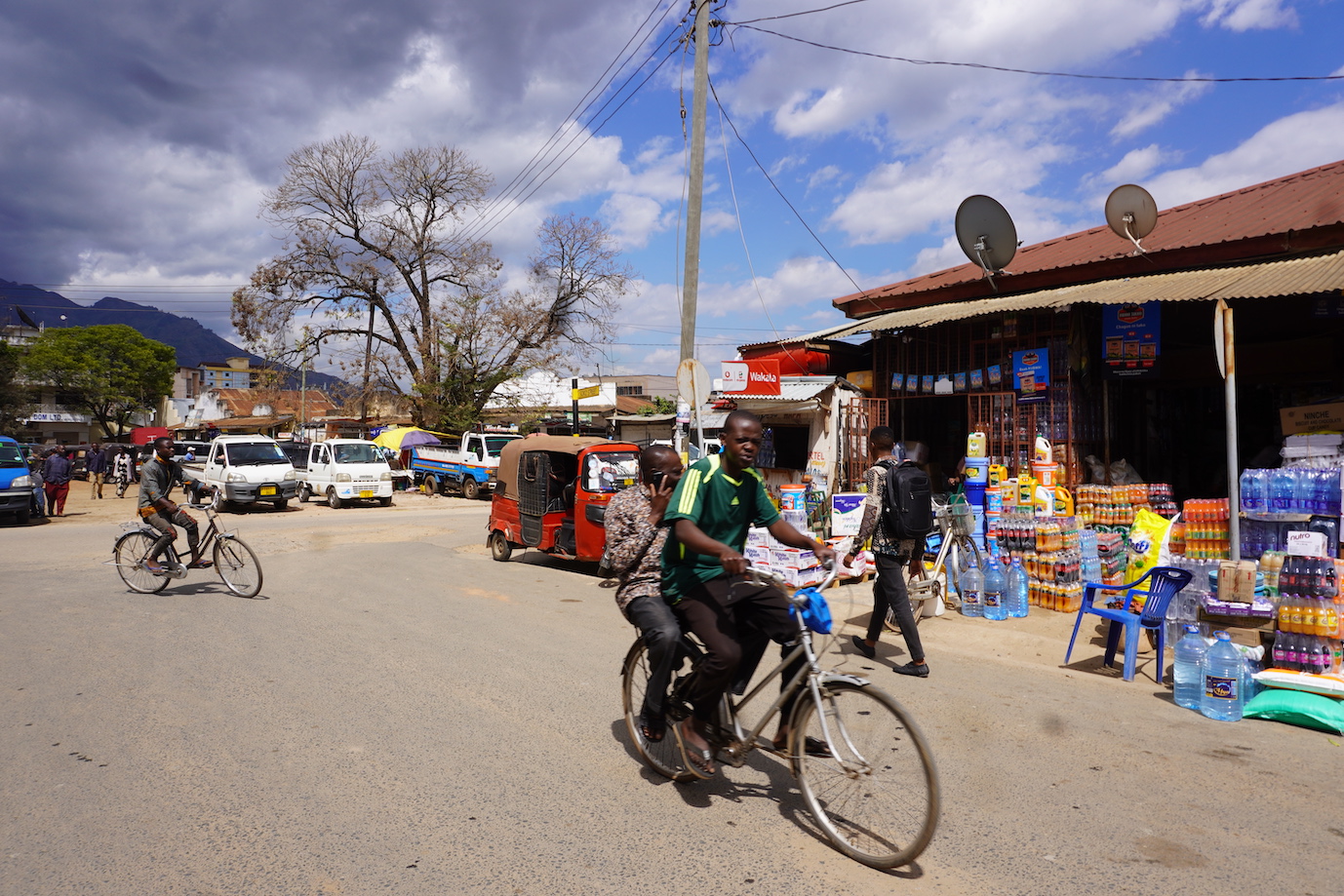 A view of the Morogoro streets. Some shops and a view of a red Tuk Tuk with a shop and some local people biking