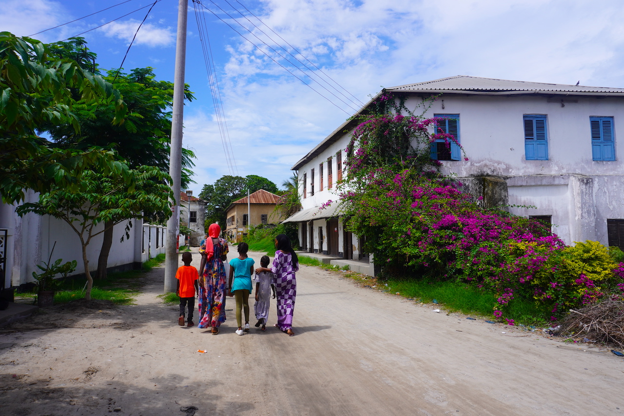 A street view of Bagamoo with 5 people walking from the back and a building with fucsia bongunvila flowers