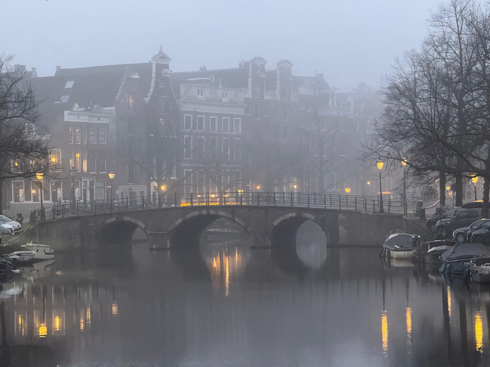 Misty winter morning over an Amsterdam canal with a historic arched bridge, canal houses, and warm streetlights reflected in the water.